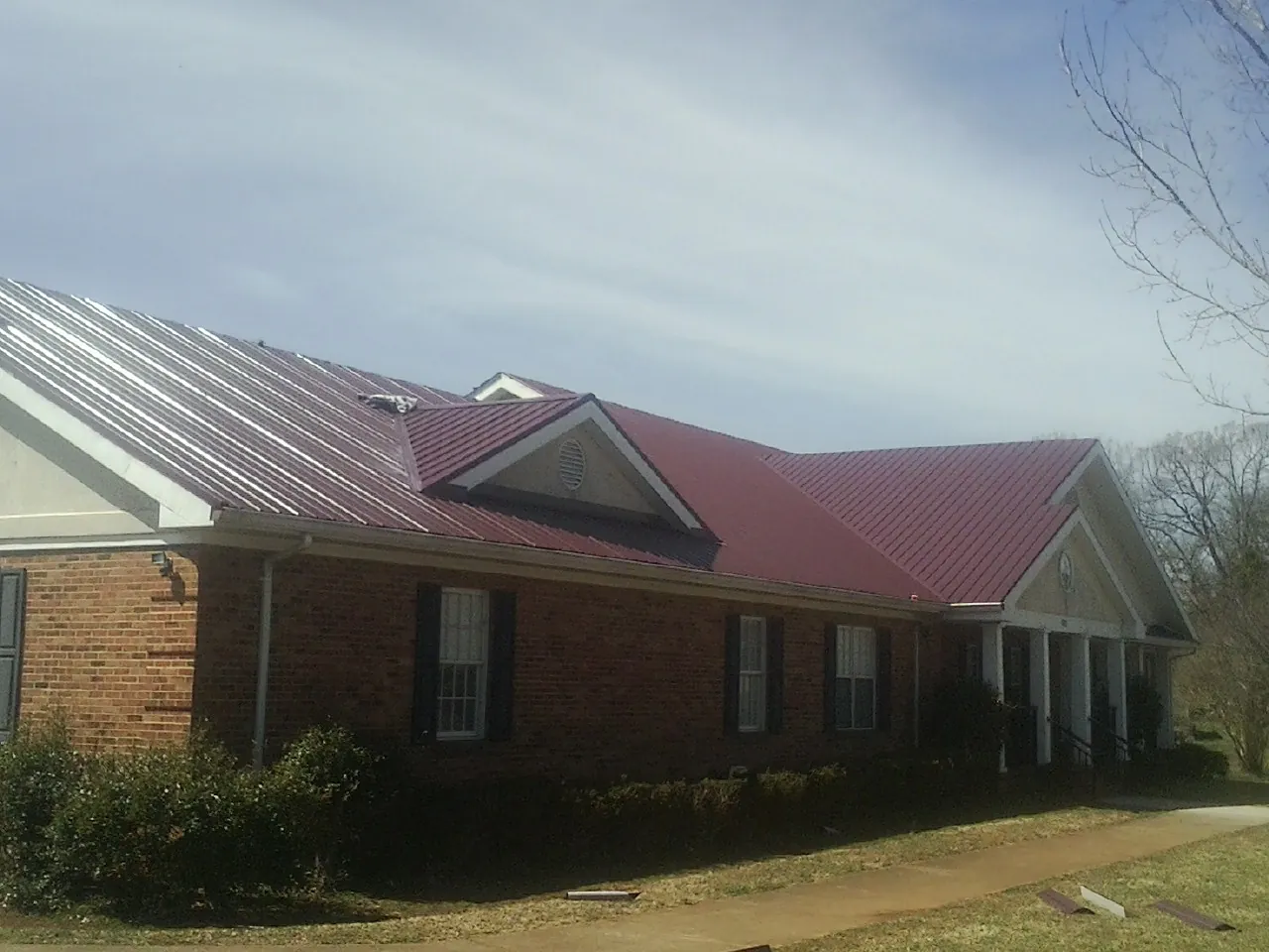 Red metal roof with gabled entries on a long brick building, from a Hail Damage Roof Repair job in Sims