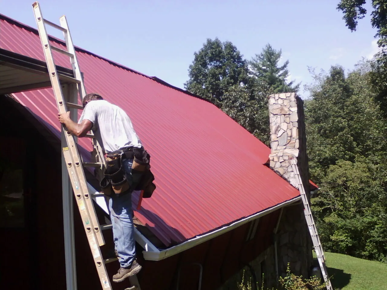 Roofer on a ladder beside a red metal roof and stone chimney — Soffit & Fascia Repair by Roofing Brothers in Sims